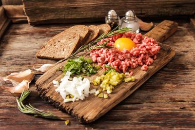 Alle schmackhaften Zutaten für Beef Tartar auf einem Holzbrett angerichtet.