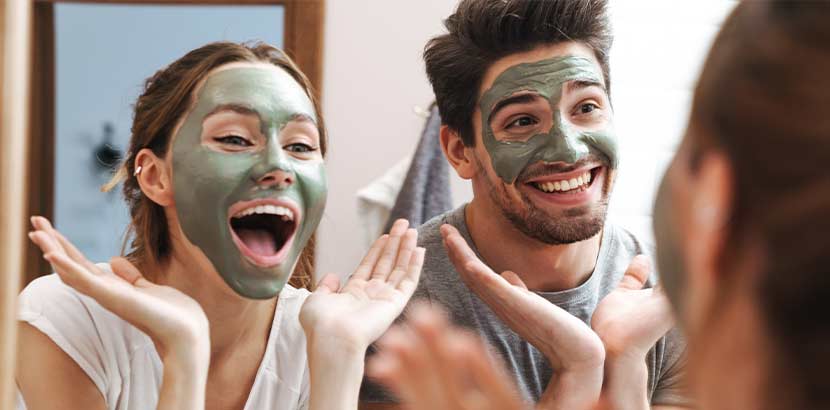 Mann und Frau mit grüner Gesichtsmaske, die vor dem Spiegel stehen und glücklich aussehen.