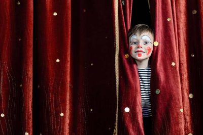 Kleiner Junge mit geschminktem Clownsgesicht, der im Kindertheater Wien durch den Vorhang auf der Bühne schaut.