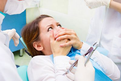Junge Frau, die im Behandlungsstuhl sitzt und sich panisch den Mund zuhält, weil sie unter Zahnarztangst und Zahnarzt Phobie leidet.