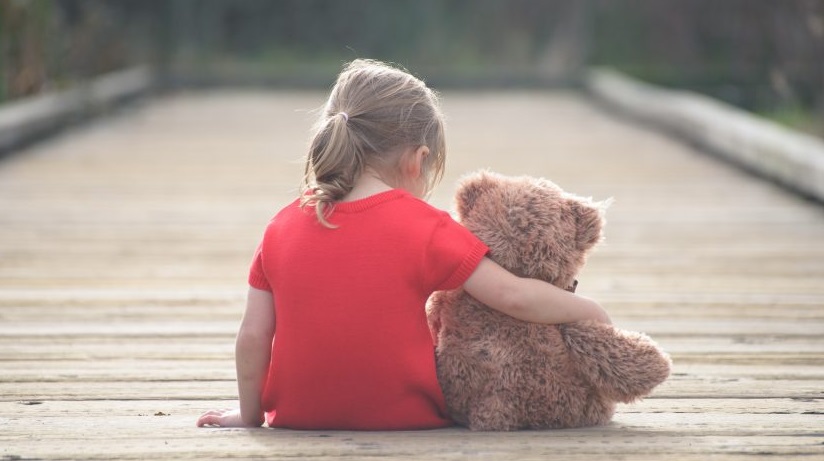 Kleines Mädchen im roten T-Shirt, das mit dem Rücken zur Kamera auf einem Steg sitzt und ihren Teddybären im Arm hält. Bestattungskosten Wien.