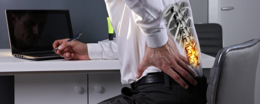 Mann im Büro, der über seinen Schreibtisch gebeugt ist und starke Schmerzen im unteren Rücken hat. Er braucht die korrekte Ergonomie am Arbeitsplatz.