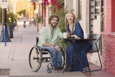 Junges Paar in einem Lokal; er sitzt im Rollstuhl. Beide lächeln die Kamera.