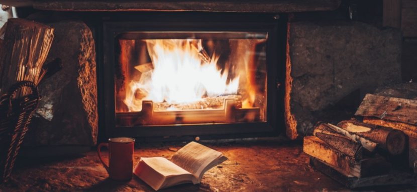 Gemütlicher Kachelofen mit Sichtfenster, vor dem ein aufgeschlagenes Buch und eine Tasse Tee stehen. Kennst du dich mit Kachelofen Preisen aus?