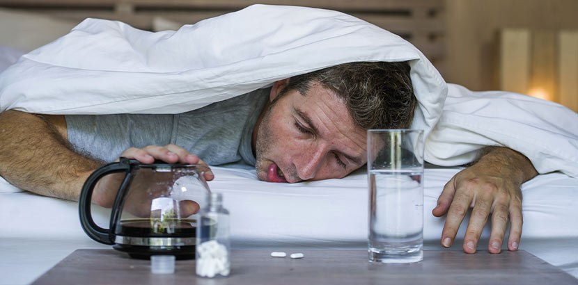Mann mit Kater, der die Bettdecke über den Kopf gezogen hat, und vor dem Kaffee, ein Glas Wasser und Tabletten stehen. Was hilft gegen Kater?