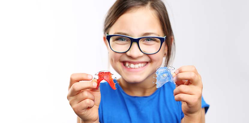 Dunkelhaariges Mädchen mit Brille, das in die Kamera lächelt und zwei verschiedene Arten von Zahnspangen hochhält.