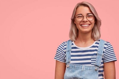 Junge blonde Frau in Latzhose und mit Zahnspange vor rosa Hintergrund.