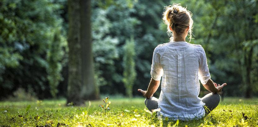 Junge Frau, die mit dem Rücken zur Kamera auf einer grünen Wiese sitzt und meditiert. Meditation Wien.