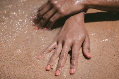 Zwei Frauenhände mit koralle-farbenen Shellac Nägeln im Sand.