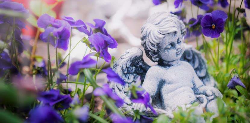 Kleiner Engel aus Gips, der am Babyfriedhof beim Zentralfriedhof Wien zwischen lila Blüten sitzt.