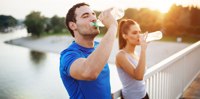 Junges Paar in Sportoutfits auf einer Brücke, beide trinken aus einer Wasserflasche. Hämorrhoiden Behandlung Wien.