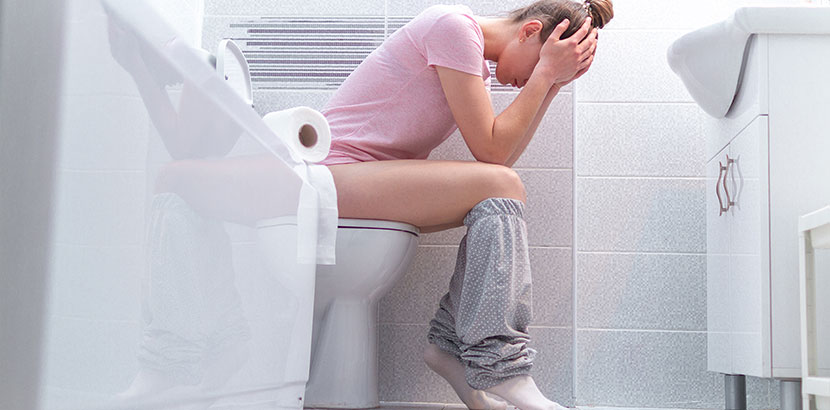 Junge Frau in rosa Shirt auf der Toilette, leidet unter Verstopfungen, hat den Kopf in die Hände gestützt. Analfissur Heilung oder OP.