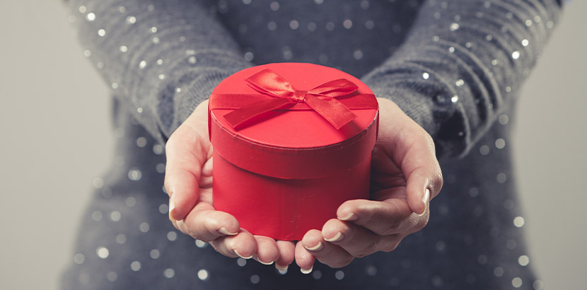 Frau im grauen Kleid, die eine rote Geschenkbox mit einem außergewöhnlichen Geschenk in die Kamera hält.