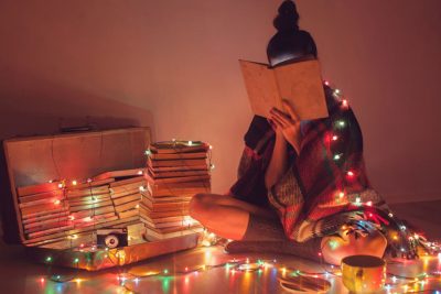 Teenager Mädchen, das sich hinter einem Buch versteckt und eine Lichterkette um sich gelegt hat. Geschenke für Teenager zu Weihnachten.
