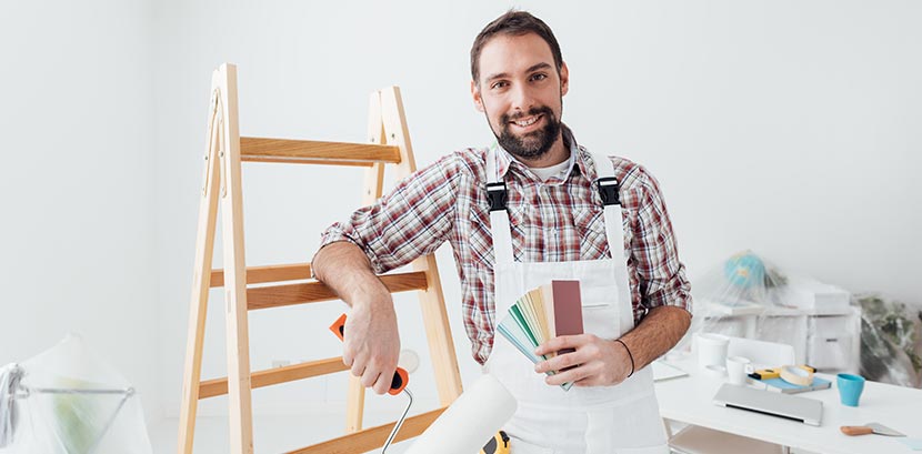 Ein Maler lehnt lächelnd an einer Leiter und hält einen Farbfächer in die Kamera