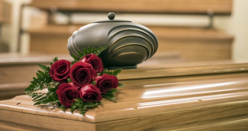 Dunkle, sparsam verzierte Urne während der Trauerfeier auf einem hellen Nussholzsarg neben roten Rosen. Feuerbestattung Wien Kosten.