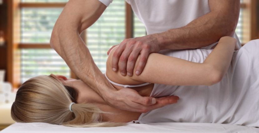 Männlicher Osteopath mit schönen starken Armen, der die Schultern und den Nacken einer jungen blonden Patientin (Rückenansicht) behandelt. Osteopathie Graz.