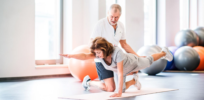 Grauhaariger Physiotherapeut, der eine ältere Patientin bei der Gymnastik unterstützt. Physiotherapie Linz.