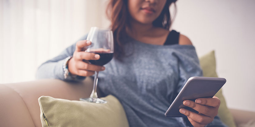 Sofa reinigen: Eine Frau trink ein Glas Rotwein auf einem Sofa und schaut auf ihr Tablet.