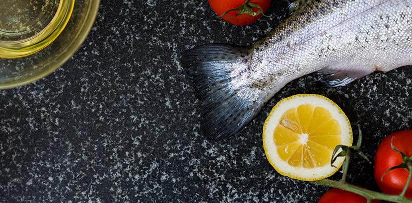 Eine dunkle Platte aus Granit, auf der eine Schale Öl, ein roher Fisch, Tomaten und eine aufgeschnittene Zitrone liegen.