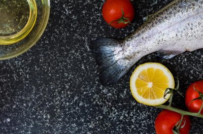 Eine dunkle Platte aus Granit, auf der eine Schale Öl, ein roher Fisch, Tomaten und eine aufgeschnittene Zitrone liegen.