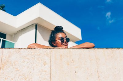 Eine Frau mit Sonnenbrille schaut hinter einer Mauer hervor