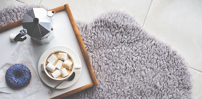 Ein Wollteppich auf Fliesen. Darauf steht ein Tablett mit einem Donut, einer Kaffeekanne und einem Schälchen Kakao und Marshmallows.