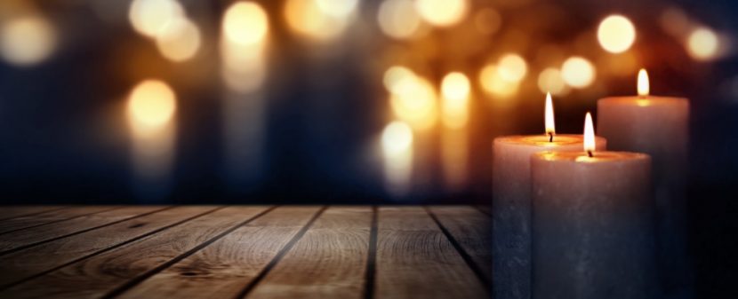Drei brennende Kerzen auf einem Holztisch vor dunklem Hintergrund, in dem noch mehr Lichter leuchten. Trauerfall Checkliste Österreich.