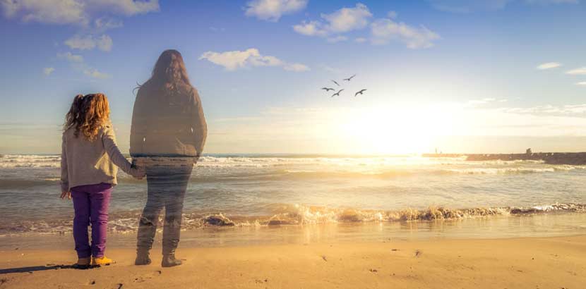 Mädchen am Strand bei Sonnenuntergang, hält die Hand ihrer verstorbenen Mutter, die ihr als Geist erscheint. Trauer Therapie Wien.