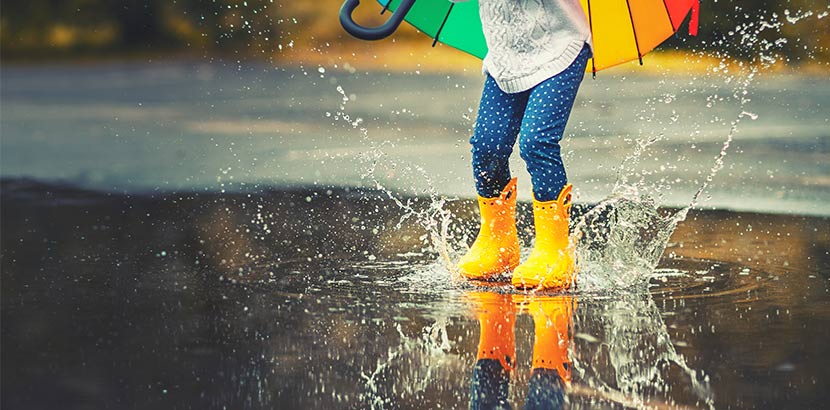 Kleines Mädchen mit buntem Schirm und gelben Gummistiefeln, das in einer Regenpfütze spielt. Zisterne einbauen.