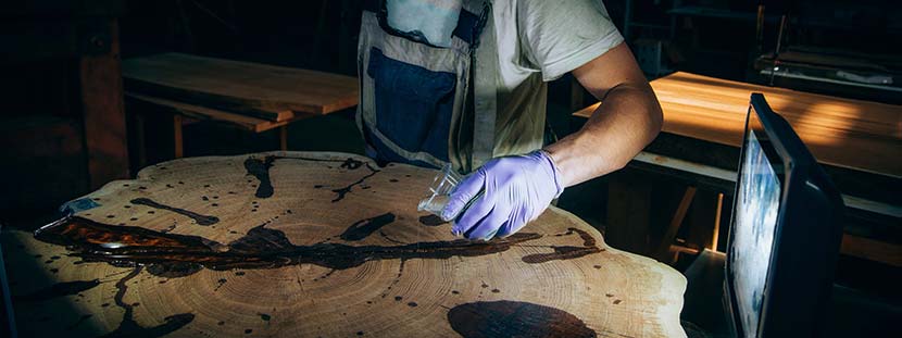 Ein Mann, der mit Epoxydharz eine Baumscheibe als Couchtisch versiegelt. Er trägt Schutzhandschuhe.