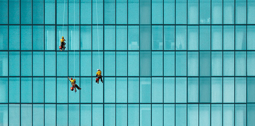Fenster putzen lassen: Drei Gebäudereiniger hängen an der Glasfassade eines großen Firmengebäudes und putzen die Fenster.