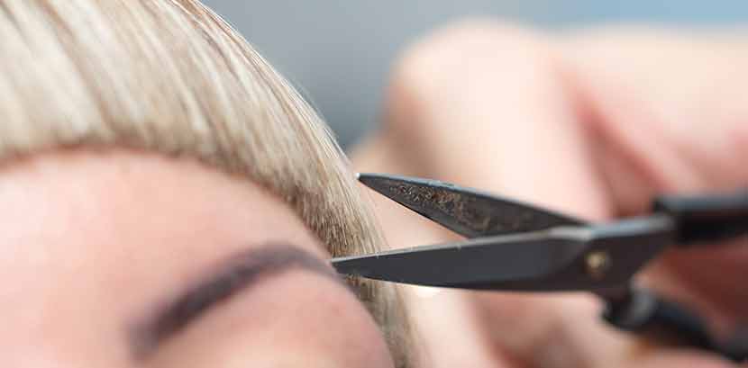 Frisurentrends 2021, Ponyfrisuren: Ein Friseur schneidet einer Dame mit blonden Haaren kurze Stirnfransen.
