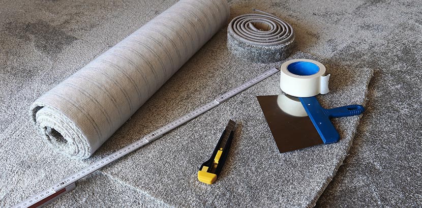 Alle Werkzeuge zum Teppich Verlegen auf einem Blick: Teppichboden, Teppichmesser, doppelseitiges Klebeband, Spachtel, Lineal und Teppichleiste