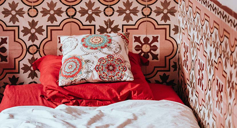 Fliesen auf Fliesen kleben: Ein Bett mit rot-weißem Bezug steht vor einer Wand mit marokkanischen Fliesen in Rot-Tönen.