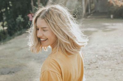 Frisurentrends 2023: Eine junge Frau mit blondem, schulterlangem Haar steht in einem sonnigen Garten und lächelt. Sie trägt ein gelbes Shirt.