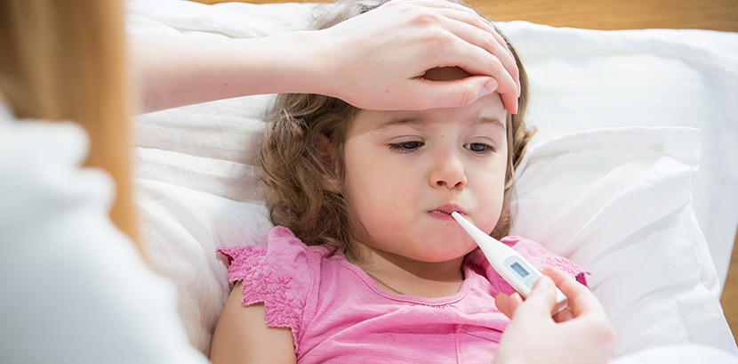 Ein Kind liegt mit Fieber durch Masern im Bett. Eine Frau misst mit einem Fieberthermometer die Temperatur des Kindes.