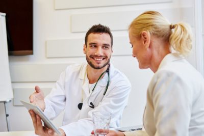 Junger Internist, der ein Gespräch mit einer blonden Patientin führt und sie dabei freundlich anlächelt. Internist Wien.