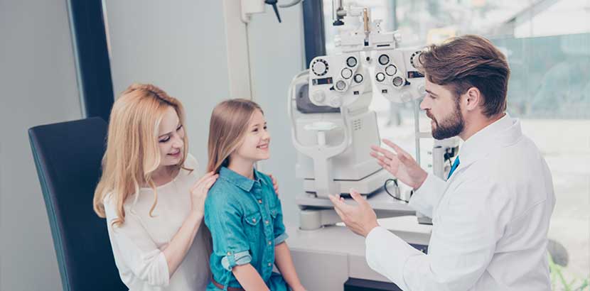 Augenarzt Wien: Kleines blondes Mädchen, das auf dem Schoß seiner Mutter sitzt, und auf die Untersuchung durch den Augenarzt wartet.