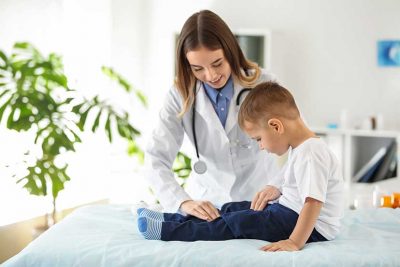 Junge Kinderorthopädin, die das Bein eines kleinen Jungen untersucht, der auf einer Behandlungsliege sitzt.