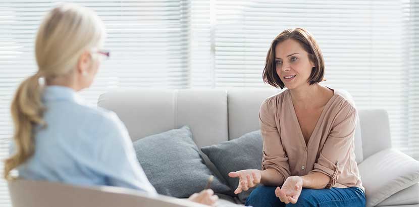 Blonde Psychotherapeutin, die mit einer jungen dunkelhaarigen Patientin spricht.