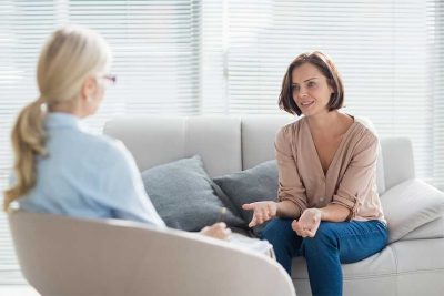 Blonde Psychotherapeutin, die mit einer jungen dunkelhaarigen Patientin spricht.