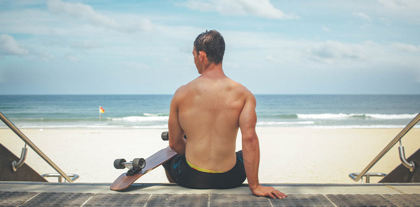 Trockene Haut durch Sonne: Ein Mann sitzt mit seinem Skateboard am Strand.