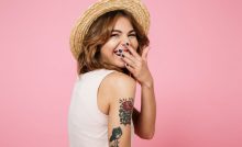 Close up portrait of a young pretty woman in summer hat laughing and covering her mouth with hand isolated over pink background