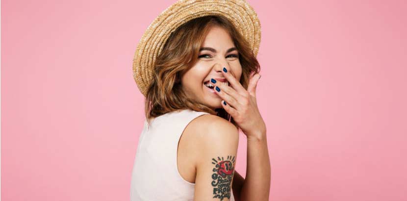 Close up portrait of a young pretty woman in summer hat laughing and covering her mouth with hand isolated over pink background