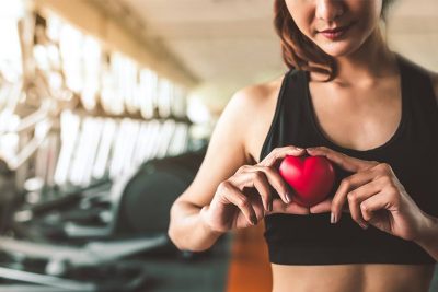 Junge dunkelhaarige Frau im Fitnessstudio, die ein kleines rotes Herz vor ihren Oberkörper hält. Sportmediziner Wien.