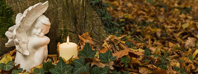 Kleiner weißer Engel neben einer brennenden weißen Kerze an einem herbstlichen Grab. Bestatter Wien.