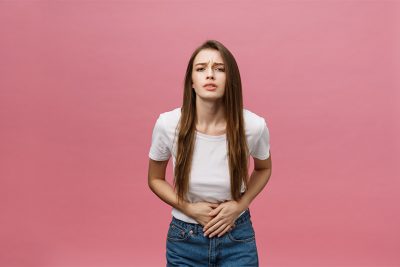 Junge Frau vor rosa Hintergund, die sich den Unterleib wegen Endometriose Schmerzen hält. Endometriose Symptome.