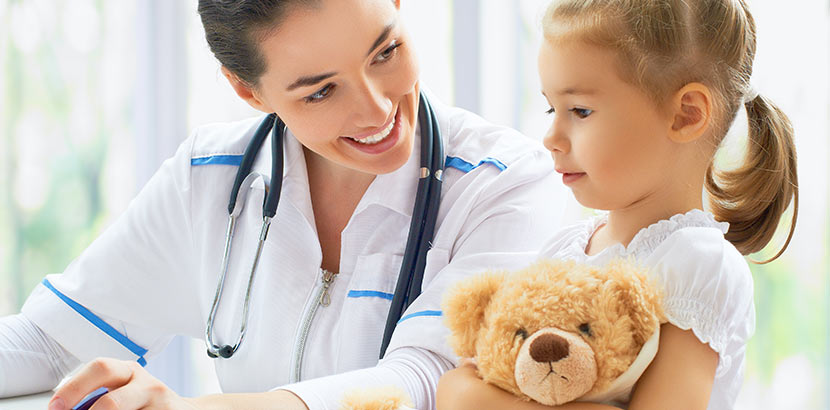 Eine Kinderärztin spricht mit einem jungen Mädchen, das einen Teddybären festhält.