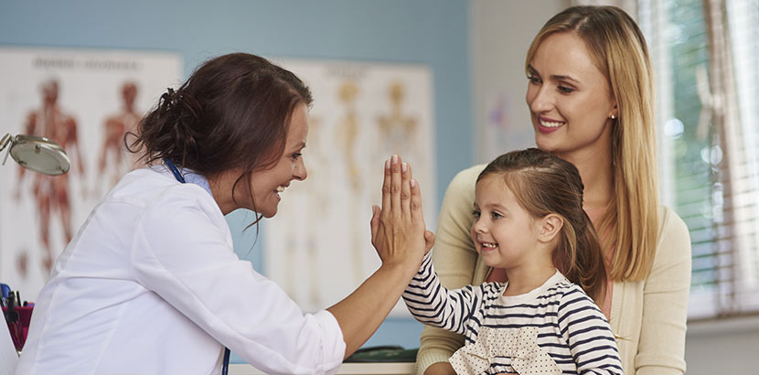 Ein junges Mädchen sitzt auf dem Schoß ihrer Mutter und gibt der Kinderärztin ein High Five. Kinderarzt Wien.